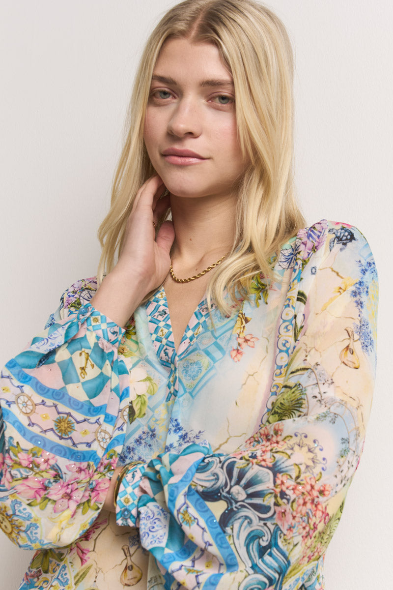 Eine junge Frau mit langen blonden Haaren trägt eine durchsichtige, fließende Princess Goes Hollywood Seidenbluse mit einem bunten Blumen- und geometrischen Muster. Sie posiert mit einer Hand im Nacken und schaut sanft in die Kamera vor einem weißen Hintergrund.