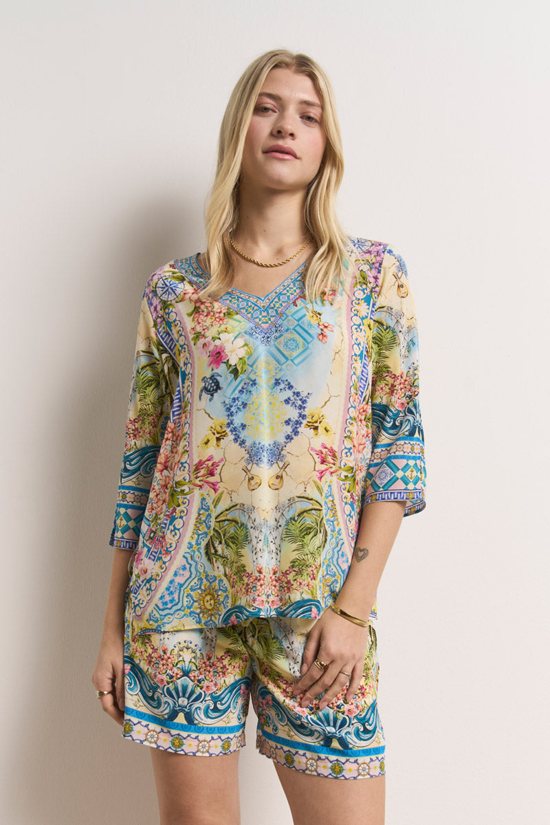 Eine blonde Frau modelliert ein farbenfrohes Set von Princess Goes Hollywood, bestehend aus einem lockeren Oberteil mit strassverzierten Bindebändern und Seidenshorts mit floralen und geometrischen Prints, und posiert vor einer schlichten weißen Wand.