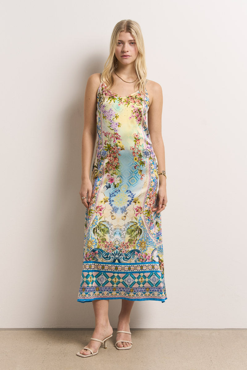 Eine Frau mit langen blonden Haaren steht vor einer schlichten Wand und trägt ein ärmelloses, knöchellanges Princess Goes Hollywood Seiden Trägerkleid mit bunten floralen und geometrischen Mustern. Dazu trägt sie Sandalen, ein Armband und eine Halskette; die Arme hat sie an der Seite.