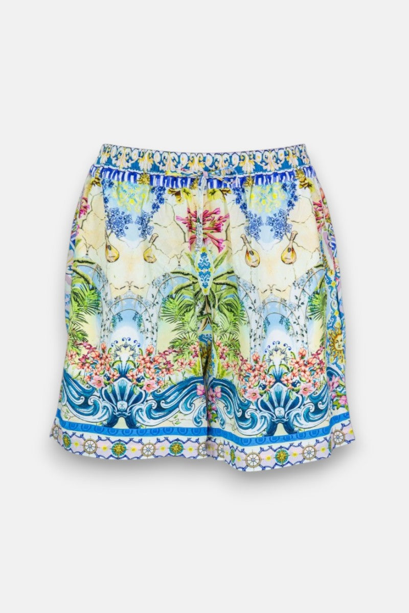Lebendige Seidenshorts von Princess Goes Hollywood mit einem tropischen Landschaftsdruck - grüne Palmen, blauer Himmel, gelbe Zitronen, rosa Blumen und verschnörkelte blaue und weiße Bordüren am elastischen Bund und Saum, auf weißem Hintergrund.