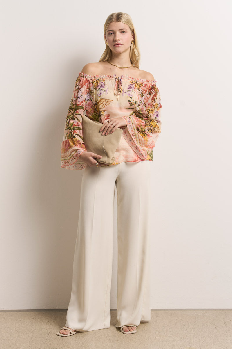 Eine Frau mit langen blonden Haaren trägt eine schulterfreie Seidenbluse mit tropischem Muster und weiten Ärmeln, weiße Hosen mit weitem Bein und offene Schuhe. Sie hält eine große beigefarbene Clutch in der Hand und steht vor einer hellen Wand.