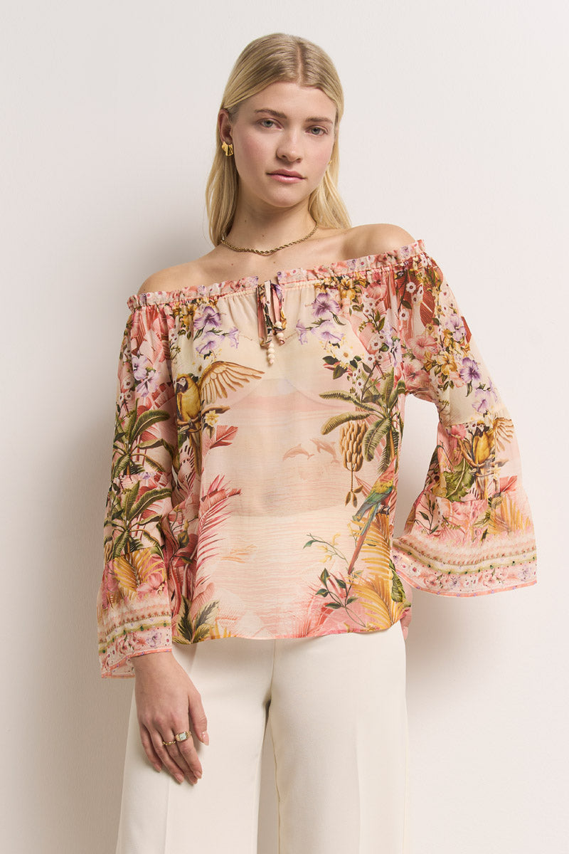 Eine Frau mit glattem blondem Haar modelliert eine schulterfreie Bluse mit tropischem Aufdruck in Rosa-, Orange- und Grüntönen mit langen Ärmeln und einer cremefarbenen Hose von Princess Goes Hollywood. Sie steht ruhig vor einer cremefarbenen Wand und blickt in die Kamera.