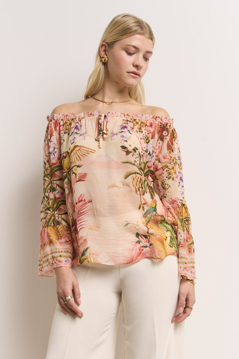 Eine blonde Frau mit goldenen Ohrringen trägt eine Carmenbluse aus Seide von Princess Goes Hollywood mit einem schulterfreien, langärmeligen tropischen Aufdruck von Blättern und Vögeln, gepaart mit einer weiten, hoch taillierten weißen Hose vor einem schlichten, hellen Hintergrund.