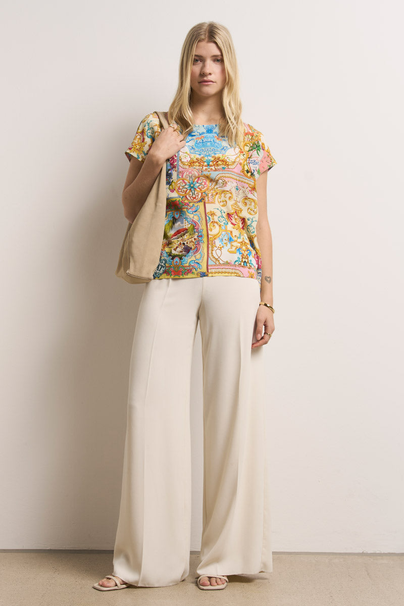 Eine Frau mit langen blonden Haaren steht vor einer schlichten hellen Wand und trägt eine bunte Bluse mit Princess Goes Hollywood-Aufdruck, eine cremefarbene Hose mit weitem Bein, Sandalen, eine beigefarbene Umhängetasche und minimalen Schmuck für einen entspannten Look.