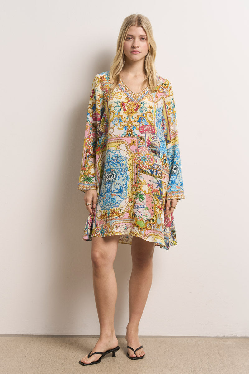 Eine Frau mit langen blonden Haaren steht vor einer weißen Wand und trägt ein lockeres Princess Goes Hollywood Blusenkleid aus Seide mit leuchtenden blauen, rosa und goldenen Motiven und einem V-Ausschnitt. Sie hat einen neutralen Gesichtsausdruck und trägt schwarze, niedrighackige Sandalen.