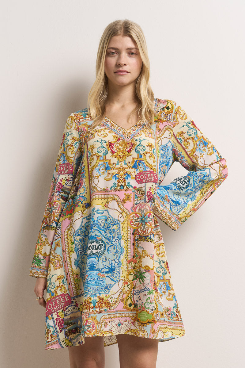 Eine Frau mit glattem blondem Haar trägt ein farbenfrohes Princess Goes Hollywood Dolce Vita-Print Blusenkleid Seide. Das langärmelige Kleid in A-Linie hat verschnörkelte blaue, gelbe, rosa und rote Muster mit "COFFEE" und "CHOCOLAT" und einen schmeichelhaften V-Ausschnitt.