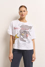 Eine Frau mit heller Haut und zurückgebundenen braunen Haaren trägt goldene Ohrringe und ein weißes Princess Goes Hollywood Tom und Jerry Baumwoll-T-Shirt, gepaart mit einer weiten blauen Hose. Auf dem T-Shirt sind Tom und Jerry abgebildet, während sie vor einer schlichten hellen Wand steht.