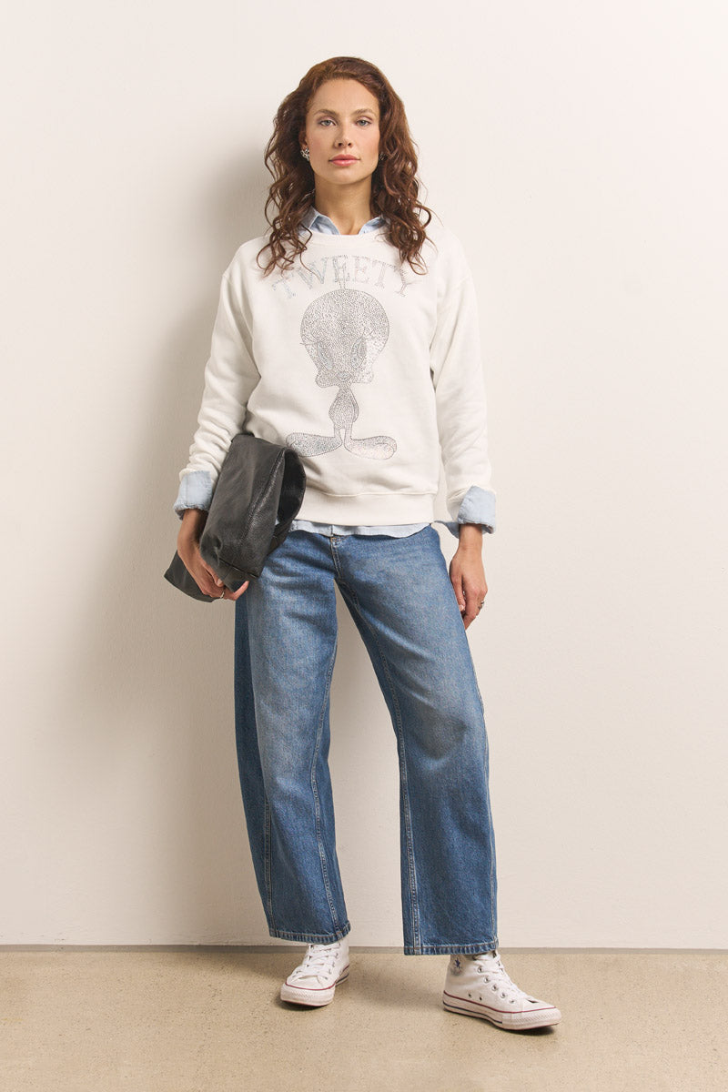 Eine Frau mit gewellten roten Haaren steht vor einer weißen Wand und trägt ein weißes Princess Goes Hollywood Tweety Sweatshirt aus Baumwolle, blaue Jeans mit Bündchen und weiße Turnschuhe. Sie hält eine große graue Clutch in der Hand und schaut mit entspanntem Blick in die Kamera.