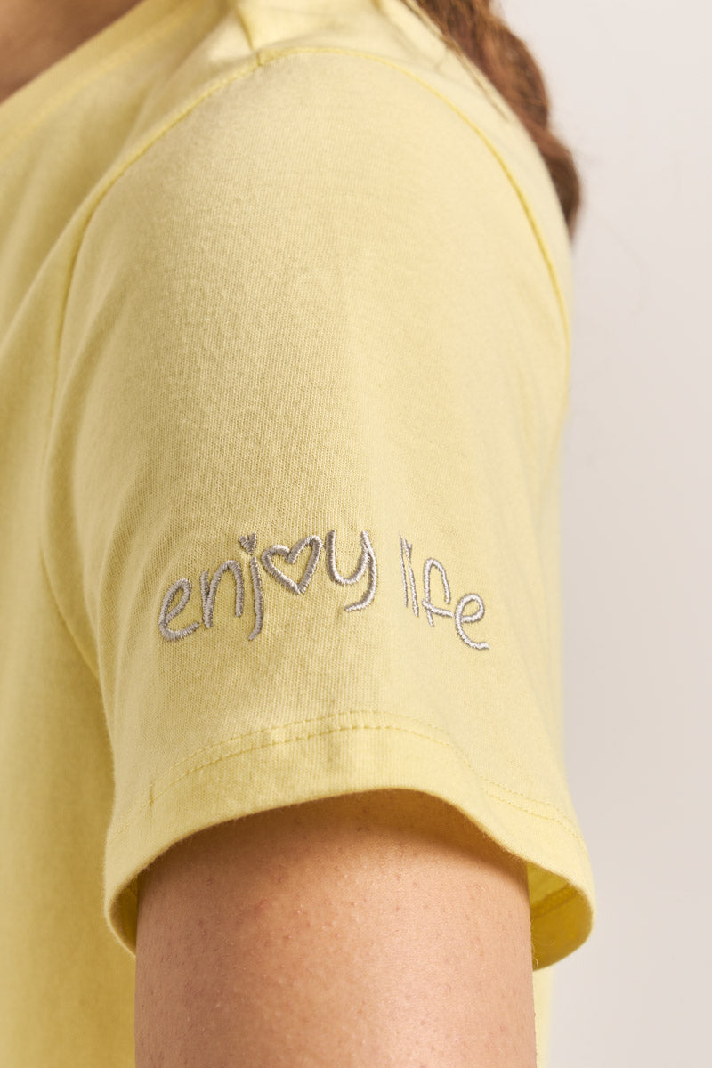 Nahaufnahme des Oberarms einer Person in einem kurzärmeligen gelben Princess Goes Hollywood Tweety-T-Shirt aus Baumwolle. Die hellgraue Stickerei auf dem Ärmel lautet "enj♥y life", wobei das "o" in "enjoy" durch ein Herzsymbol ersetzt ist.