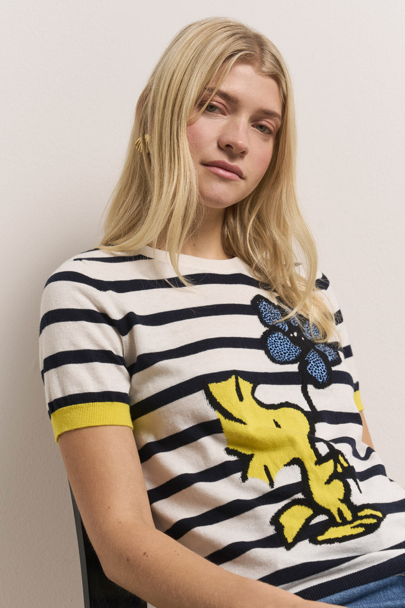 Eine junge Frau mit langen blonden Haaren sitzt an einer schlichten Wand und trägt einen weißen Pullover aus einer Baumwoll-Kaschmir-Mischung mit schwarzen Streifen, gelben Manschetten und einem großen gelben Cartoon-Hund, der eine blaue Blume auf der Vorderseite hält.