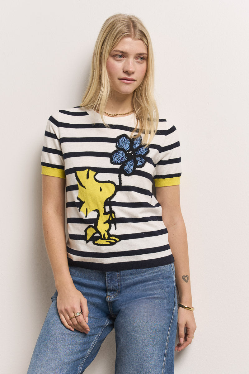 Eine Frau mit langen blonden Haaren trägt einen gestreiften Pullover aus Baumwoll-Kaschmir-Gemisch von Princess Goes Hollywood mit einem großen Woodstock-Motiv und gelben Akzenten. Sie trägt dazu Blue Jeans und Ringe und steht vor einer weißen Wand.