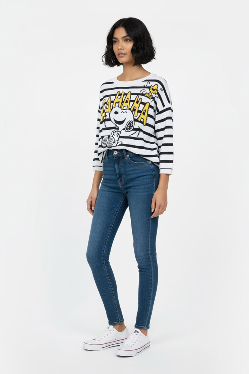 Eine Frau mit kinnlangen dunklen Haaren trägt einen Princess Goes Hollywood gestreifter Pullover mit Snoopy-Motiv, blaue Skinny Jeans und weiße Turnschuhe. Sie steht vor einem schlichten weißen Hintergrund und schaut mit neutralem Blick leicht nach links.