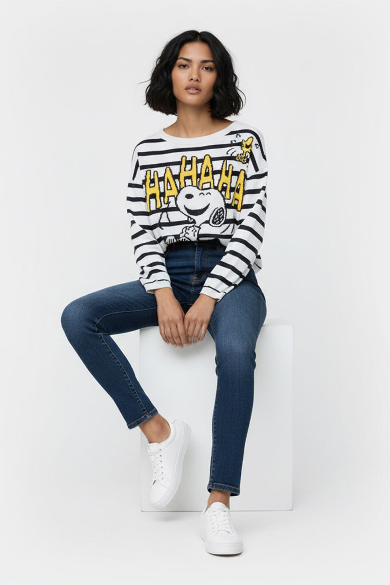 Eine Frau mit gewelltem Bob sitzt auf einem weißen Würfel in blauen Jeans, weißen Turnschuhen und einem gestreiften Snoopy & Woodstock-Pullover von Princess Goes Hollywood mit fettgedrucktem gelben "HA HA HA"-Schriftzug aus einer weichen Baumwollmischung.