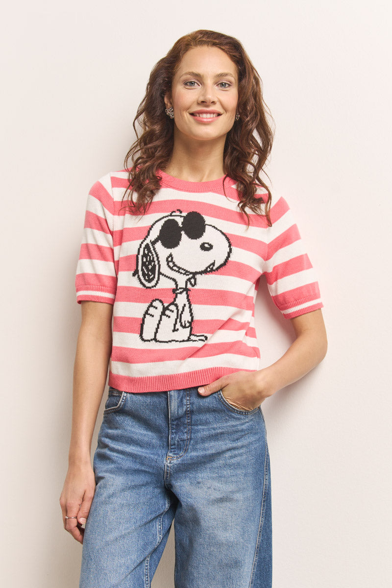 Eine Frau mit gewelltem braunem Haar lächelt in die Kamera. Sie trägt einen rosa-weiß gestreiften Baumwollmischpullover mit Snoopy-Motiv und hellblaue Jeans mit hoher Taille und steht mit einer Hand in der Tasche vor einer hellen Wand.