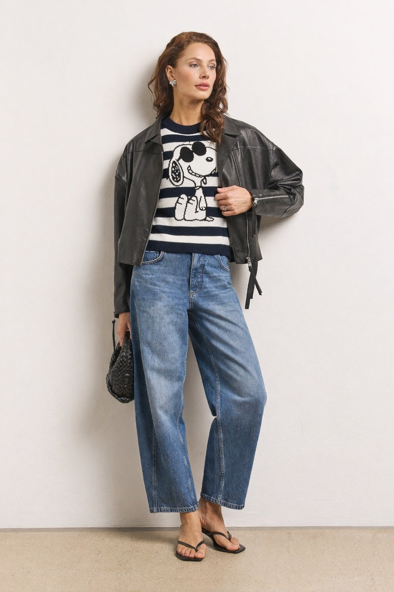 Eine Frau steht vor einer weißen Wand und trägt lockere Bluejeans, schwarze Flip-Flops und einen Gestreifter Pullover mit Snoopy-Motiv von Princess Goes Hollywood. Sie trägt eine schwarze, strukturierte Tasche und ihre gewellten braunen Haare sind zu einer Seite gestylt.