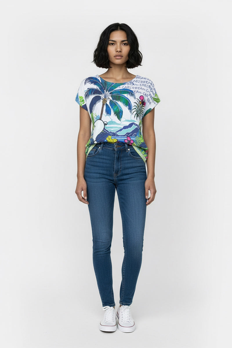 Eine Frau mit kurzen schwarzen Haaren blickt nach vorne. Sie trägt ein farbenfrohes Bio-Baumwolle-T-Shirt mit Palmen, Vögeln und einem verspielten Snoopy-Aufdruck, blaue High-Waisted-Skinny-Jeans und weiße Turnschuhe vor einem schlichten weißen Hintergrund.