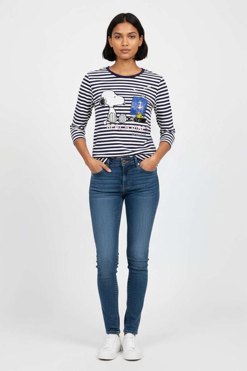 Eine Frau mit glattem dunklem Haar und neutralem Gesichtsausdruck blickt nach vorne. Sie trägt ein marineblau-weiß gestreiftes Baumwollhemd mit Snoopy-Aufdruck von Princess Goes Hollywood, taillierte Bluejeans und weiße Turnschuhe. Hände in den Taschen. Einfarbig weißer Hintergrund.