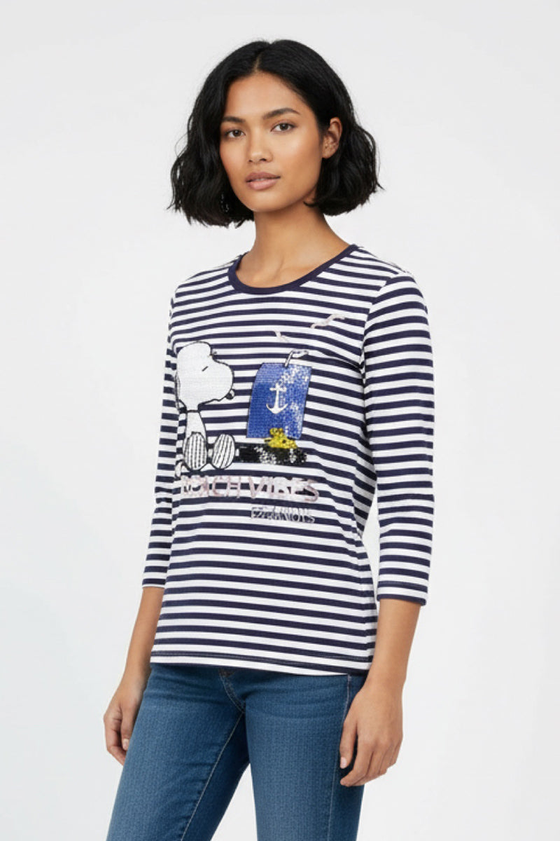 Eine Frau mit kurzen, gewellten schwarzen Haaren trägt ein marineblau-weiß gestreiftes Baumwollhemd von Princess Goes Hollywood mit Snoopy- und maritimen Motiven, kombiniert mit blauen Jeans vor einem schlichten hellen Hintergrund.