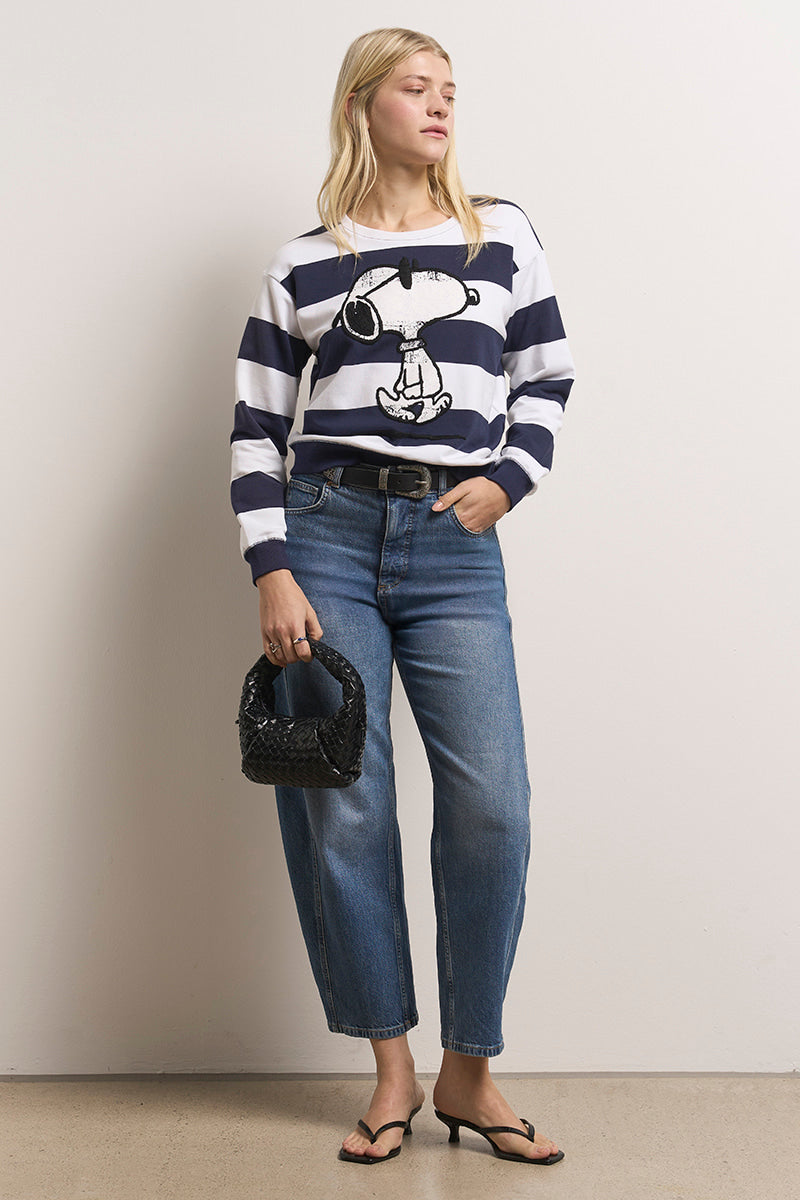 Eine Frau mit langen blonden Haaren steht an einer schlichten Wand und trägt ein gestreiftes Baumwollmisch-Sweatshirt mit Snoopy-Aufdruck, lässige Bluejeans, schwarze Flip-Flops und eine kleine schwarze Stofftasche und schaut ruhig zur Seite.