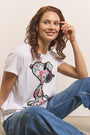 Eine Frau mit gewelltem kastanienbraunem Haar und heller Haut sitzt lässig lächelnd da. Sie trägt ein Princess Goes Hollywood weißes Baumwolle Snoopy T-Shirt mit buntem Blumenprint und lässige Blue Jeans. Ihre Pose ist lässig vor einem neutralen Hintergrund.