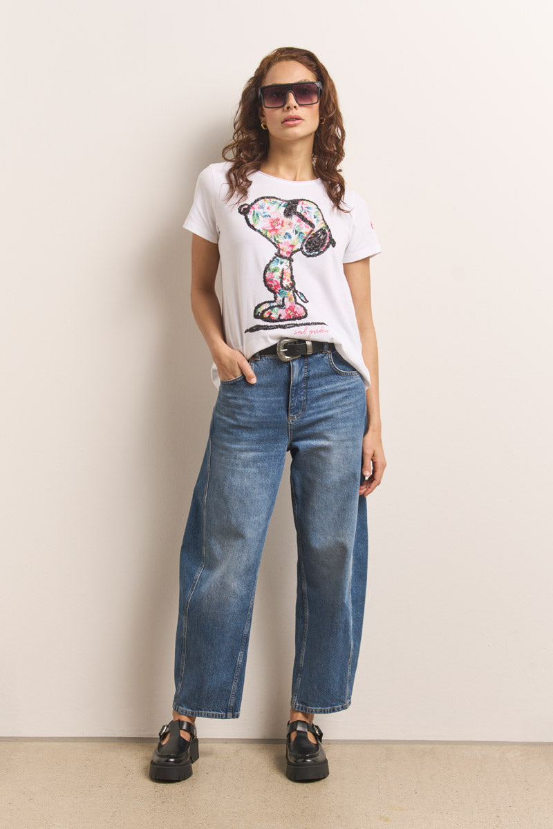 Eine Frau mit gewelltem braunem Haar trägt eine übergroße Sonnenbrille, weite blaue Jeans, schwarze klobige Sandalen und ein Princess Goes Hollywood Snoopy-T-Shirt mit einer bunten Trophäengrafik. Sie posiert vor einer schlichten hellen Wand und hat eine Hand in ihrer Tasche.