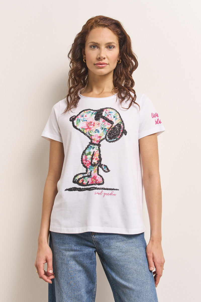 Eine Frau mit langen, gewellten kastanienbraunen Haaren trägt ein weißes Princess Goes Hollywood Snoopy T-Shirt aus Baumwolle mit einem großen, bunten Blumenprint und einem rosa "live life" Text auf dem Ärmel. Dazu trägt sie eine blaue Jeans vor einem hellen Hintergrund.