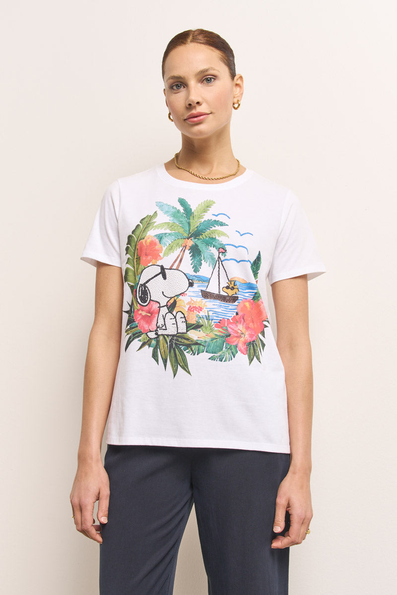 Eine Frau mit hochgesteckten braunen Haaren trägt ein Princess Goes Hollywood Snoopy T-Shirt mit einem bunten Aufdruck von tropischen Blumen, Palmen und einem Segelboot. Mit goldenen Ohrringen, einer Halskette und einer dunklen Hose steht sie ruhig vor einer hellen Wand.