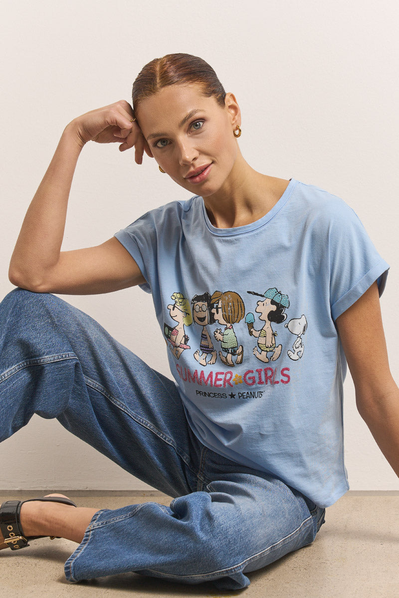 Eine Frau mit kastanienbraunem Haar sitzt vor einer weißen Wand und lächelt sanft. Sie trägt goldene Ohrringe, blaue Jeans, schwarze Schuhe und ein hellblaues Princess Goes Hollywood Baumwolle T-Shirt mit Peanuts-Aufdruck und dem Text "SUMMER GIRLS".