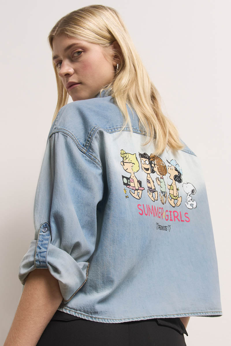 Eine blonde Frau mit glattem Haar blickt über ihre Schulter und trägt ein hellblaues Princess Goes Hollywood Jeanshemd aus Baumwolle. Auf der Rückseite sind Peanuts-Cartoonfiguren in Badeanzügen und der rosa Text "SUMMER GIRLS" über einem kleineren Schriftzug zu sehen. Die Ärmel sind teilweise aufgerollt.
