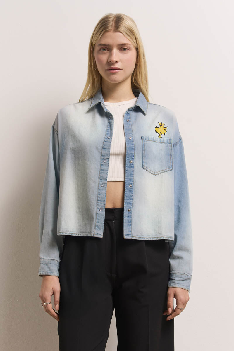 Eine junge Frau mit glattem blondem Haar steht vor einer hellen Wand. Sie trägt ein Jeanshemd mit Peanuts-Print und gelbem Woodstock-Aufnäher, darüber ein weißes Crop-Top und eine schwarze Hose mit hoher Taille. Ihr Gesichtsausdruck ist neutral.