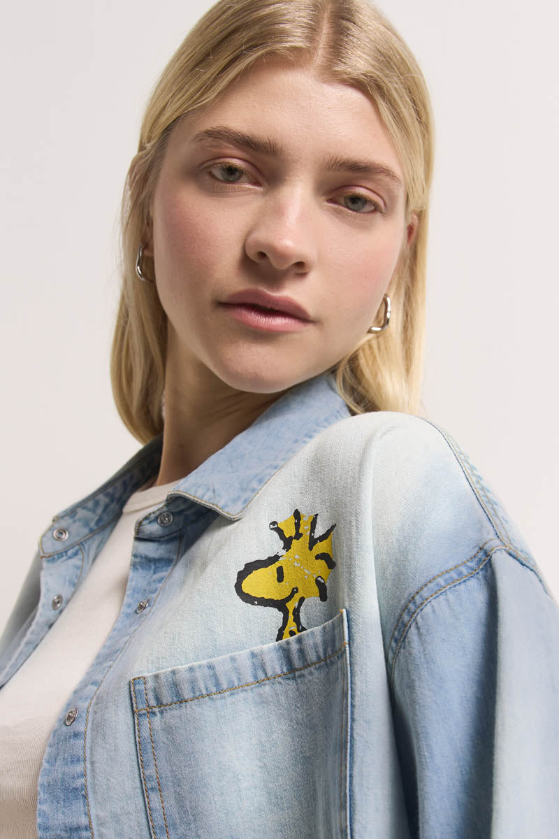 Eine junge Frau mit glattem blondem Haar trägt ein Princess Goes Hollywood Jeanshemd aus leichter Baumwolle über einem weißen Oberteil. Auf der Brusttasche ist ein gelber Cartoon-Vogel wie Woodstock aufgestickt. Sie hat einen neutralen Gesichtsausdruck und silberne Ohrringe.