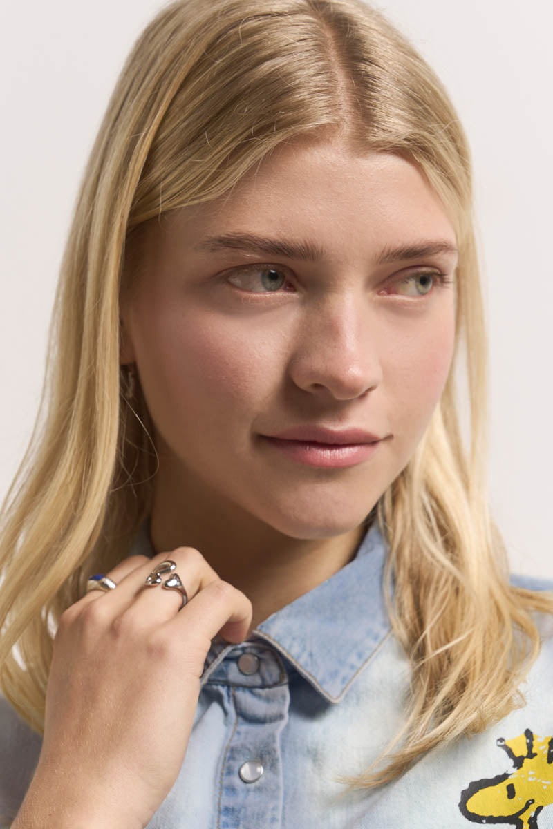 Eine junge blonde Frau schaut nach links, sie trägt ein helles Jeanshemd von Princess Goes Hollywood mit einem gelben Aufnäher. Sie hält ihren Kragen und zeigt einen silbernen Ring. Ihr ruhiger Gesichtsausdruck kontrastiert mit dem sanft beleuchteten neutralen Hintergrund.