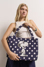 Eine junge Frau mit langen blonden Haaren trägt ein ärmelloses weißes Oberteil und einen dunklen Rock. Sie hält eine Princess Goes Hollywood navy Baumwollcanvas Tasche mit Snoopy-Stickerei, geräumig für einen 15-Zoll Laptop, und lächelt dezent vor einem weißen Hintergrund.
