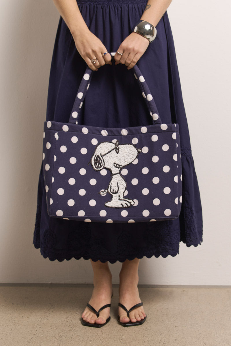 Eine Person in einem marineblauen Rock und schwarzen Flip-Flops hält eine Princess Goes Hollywood Tasche aus marineblauem Baumwollcanvas mit Snoopy-Stickerei, gestylt mit Silberschmuck vor einer schlichten hellen Wand.