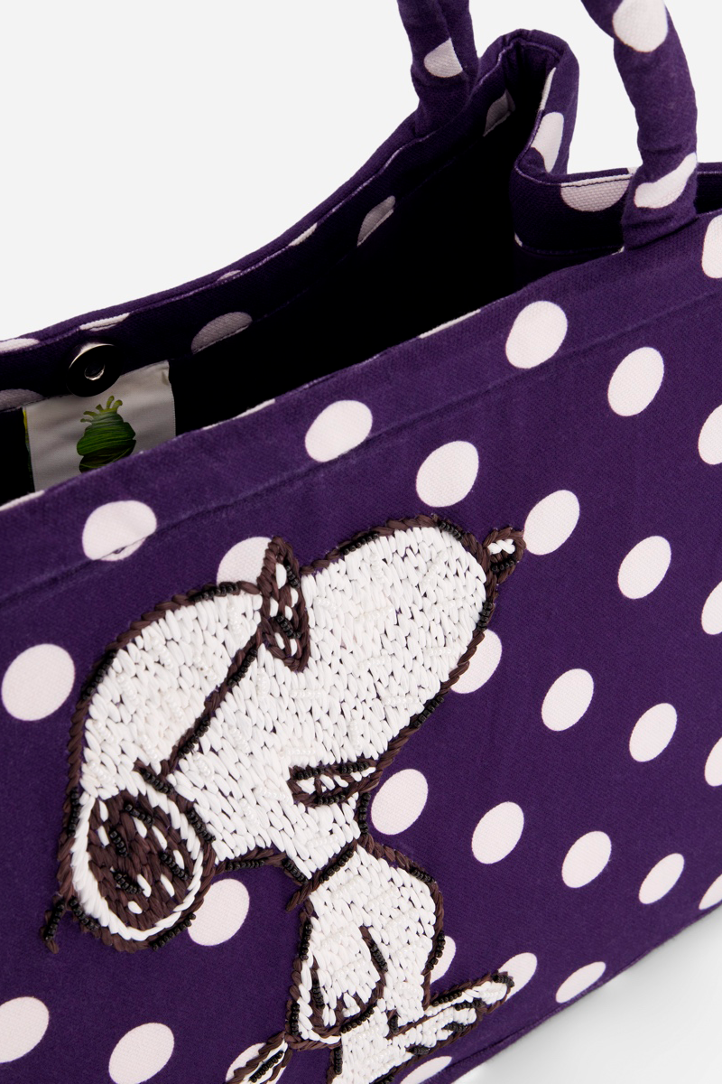 Eine Nahaufnahme einer lilafarbenen Baumwollcanvas-Tasche von Princess Goes Hollywood mit weißen Tupfen, Snoopy-Stickerei auf der Vorderseite, passend für einen 15-Zoll-Laptop, mit passenden Griffen und einem blattförmigen Anhänger im Inneren.
