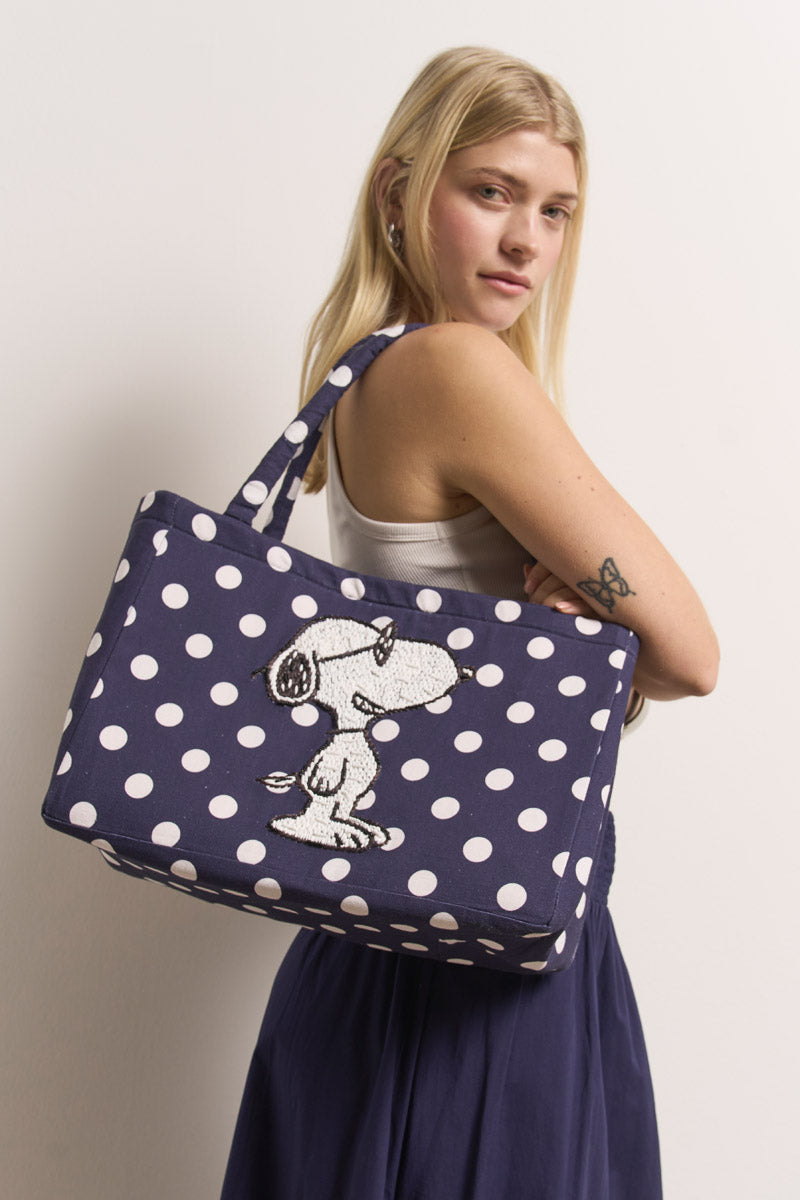 Eine blonde Frau in einem weißen Tanktop und einem marineblauen Rock posiert seitlich und hält eine Princess Goes Hollywood marineblaue Baumwollcanvas-Tasche mit Snoopy-Stickerei. Sie hat ein Schmetterlings-Tattoo auf dem Oberarm und steht vor einem schlichten weißen Hintergrund.