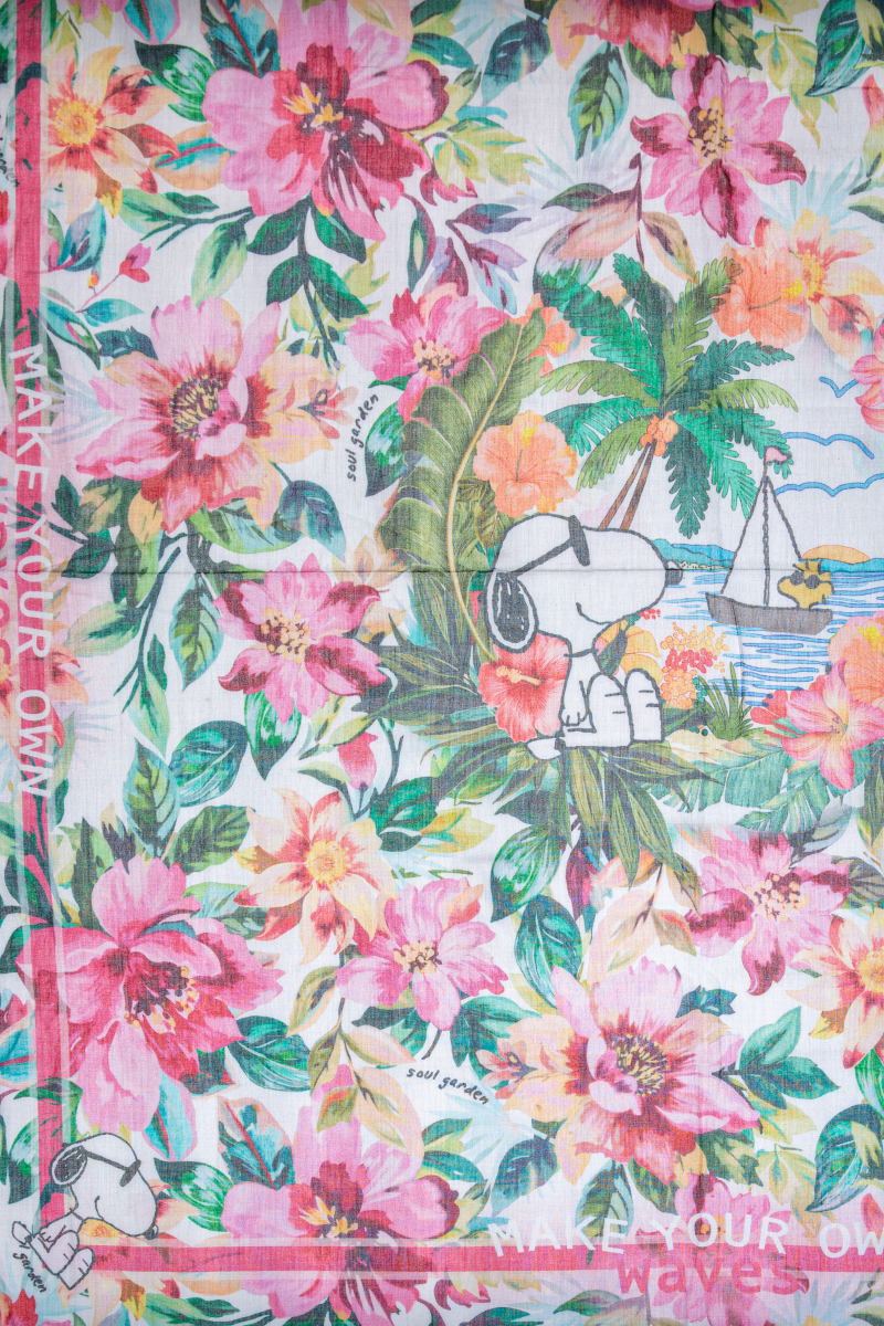 Auf dem farbenfrohen Princess Goes Hollywood-Schal aus Baumwollmodal ist Snoopy mit Sonnenbrille zwischen rosa-roten Blumen, grünen Blättern, Palmen, einem blauen Segelboot und Woodstock am Strand mit dem Slogan "MAKE YOUR OWN waves" zu sehen.