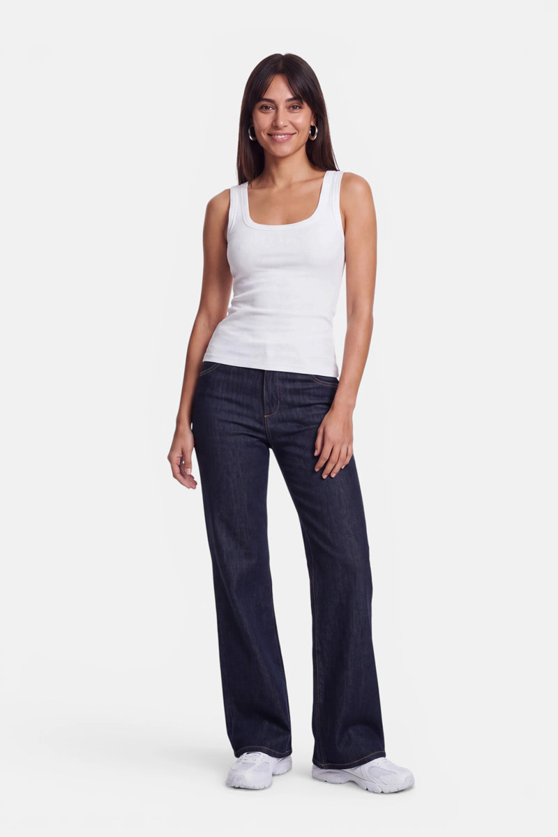 Eine Frau steht vor einem weißen Hintergrund und trägt ein weißes Basic-Top mit Rundhalsausschnitt aus weicher Baumwolle, dazu eine dunkelblaue Jeans mit geradem Bein und weiße Sneaker. Sie hat langes, glattes Haar, lächelt und hält ihren linken Arm entspannt an der Seite.