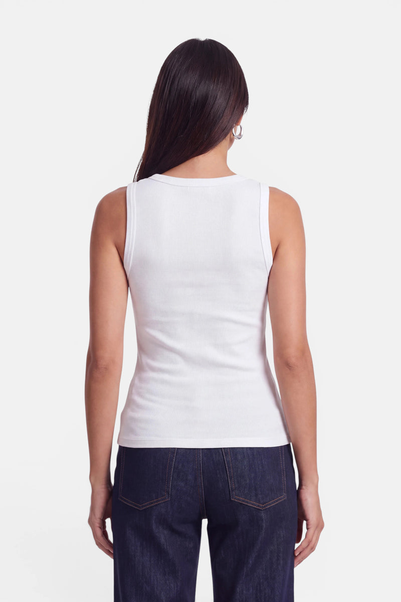 Eine Frau mit langen, dunklen Haaren steht mit dem Rücken zur Kamera und trägt ein eng anliegendes, weißes, ärmelloses Basic-Top mit Rundhalsausschnitt sowie dunkelblaue Jeans. Der Hintergrund ist schlicht weiß. Ihre Arme hängen entspannt an den Seiten herab, und sie steht aufrecht. Der Look aus weicher Baumwolle wirkt mühelos lässig und zeitlos.