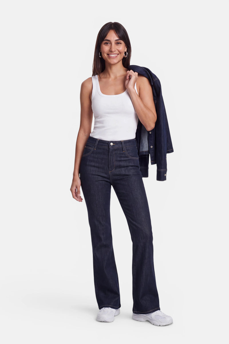 Eine Frau mit langen braunen Haaren lächelt. Sie trägt ein weißes Basic-Top aus Baumwolle mit Rundhalsausschnitt, dunkelblaue Jeans mit hoher Taille und weiße Sneaker. Sie hält eine dazu passende Jeansjacke über ihrer linken Schulter und steht vor einem schlichten weißen Hintergrund.