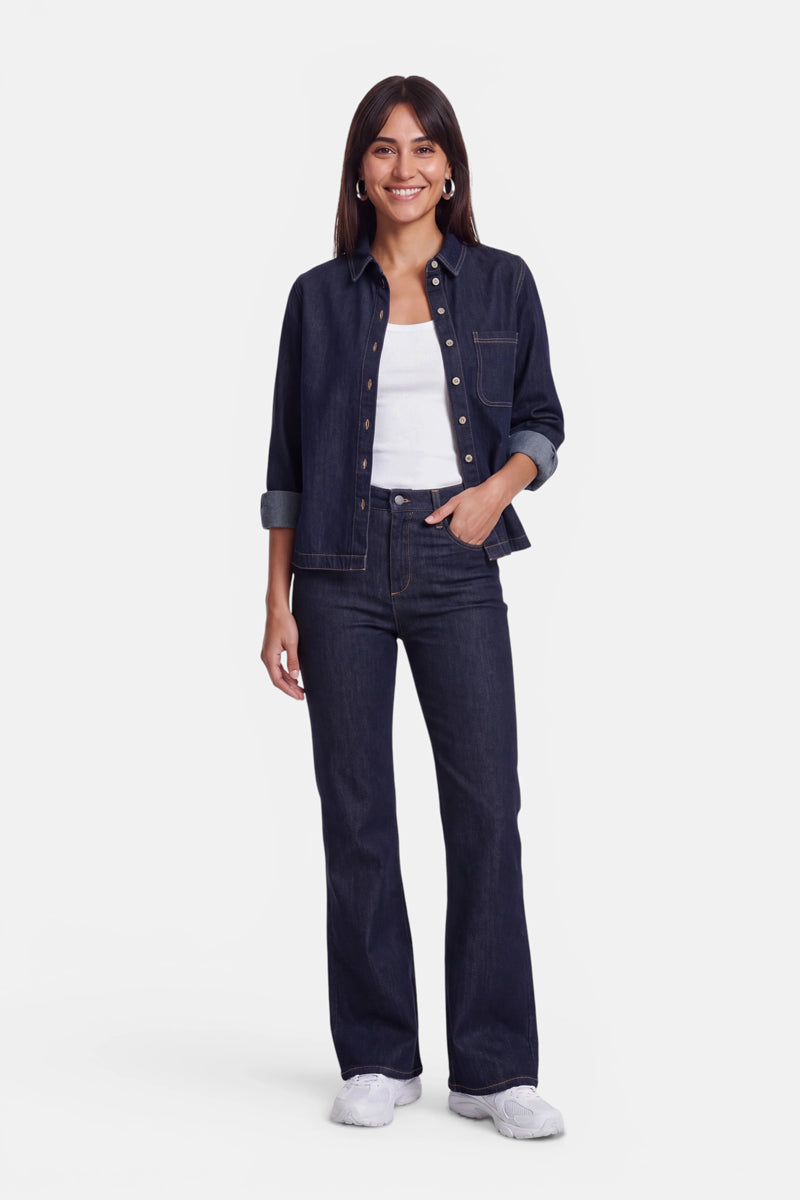 Eine Frau mit glattem braunem Haar trägt ein dunkles Jeanshemd mit Knöpfen über einem weißen Basic-Top aus Baumwolle mit Rundhalsausschnitt, dazu eine dunkle Jeans und weiße Sneaker. Sie steht lächelnd mit einer Hand in der Hosentasche vor einem schlichten weißen Hintergrund.