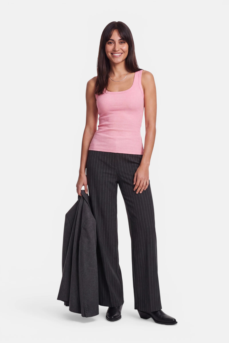 Eine Frau mit langen, dunklen Haaren trägt ein rosa Basic-Top aus Baumwolle mit Rundhalsausschnitt und eine schwarze Nadelstreifenhose; in der linken Hand hält sie einen dunklen Blazer. Sie steht vor einem schlichten weißen Hintergrund, trägt schwarze Schuhe und lächelt.