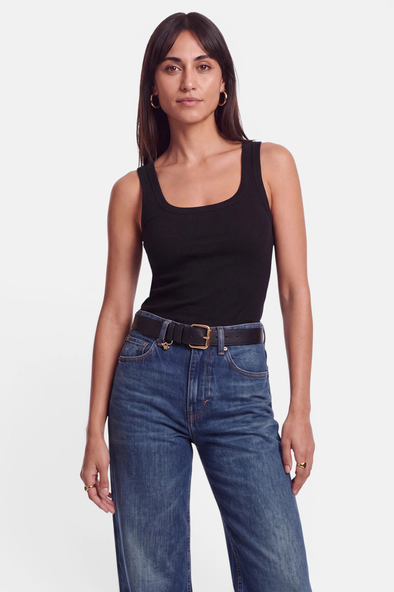 Eine Frau mit glattem, dunklem Haar trägt ein ärmelloses, schwarzes Basic-Top aus Baumwolle mit Rundhalsausschnitt, eine blaue Jeans mit hoher Taille und einen schwarzen Gürtel mit goldener Schnalle. Sie steht vor einem schlichten weißen Hintergrund und blickt mit ausdruckslosem Gesichtsausdruck in die Kamera.
