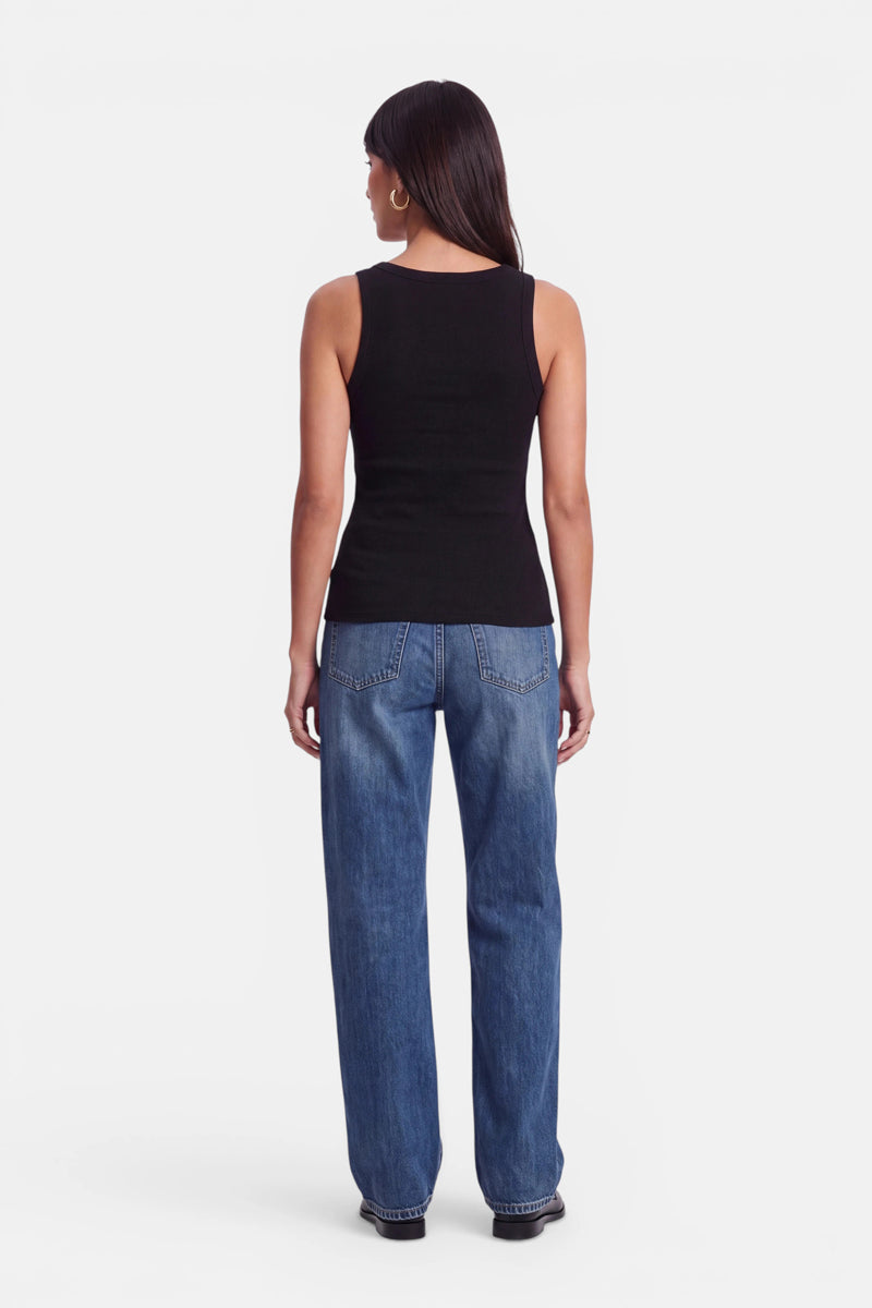Eine Frau steht mit dem Rücken zur Kamera und trägt ein schwarzes, ärmelloses Basic-Top mit Rundhalsausschnitt sowie eine lockere blaue Jeans. Sie hat glattes, dunkles Haar und trägt Creolen. Der Hintergrund ist schlicht weiß.