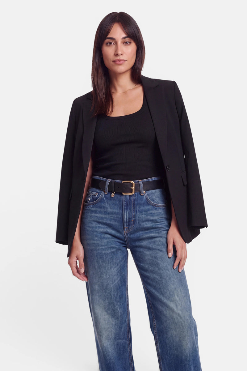 Eine Frau mit glattem, dunklem Haar trägt einen schwarzen Blazer, der ihr über die Schultern drapiert ist, ein schwarzes Basic-Oberteil mit Rundhalsausschnitt aus weicher Baumwolle sowie eine blaue Jeans mit hoher Taille und einem schwarzen Gürtel. Sie steht vor einem schlichten weißen Hintergrund und blickt selbstbewusst in die Kamera.