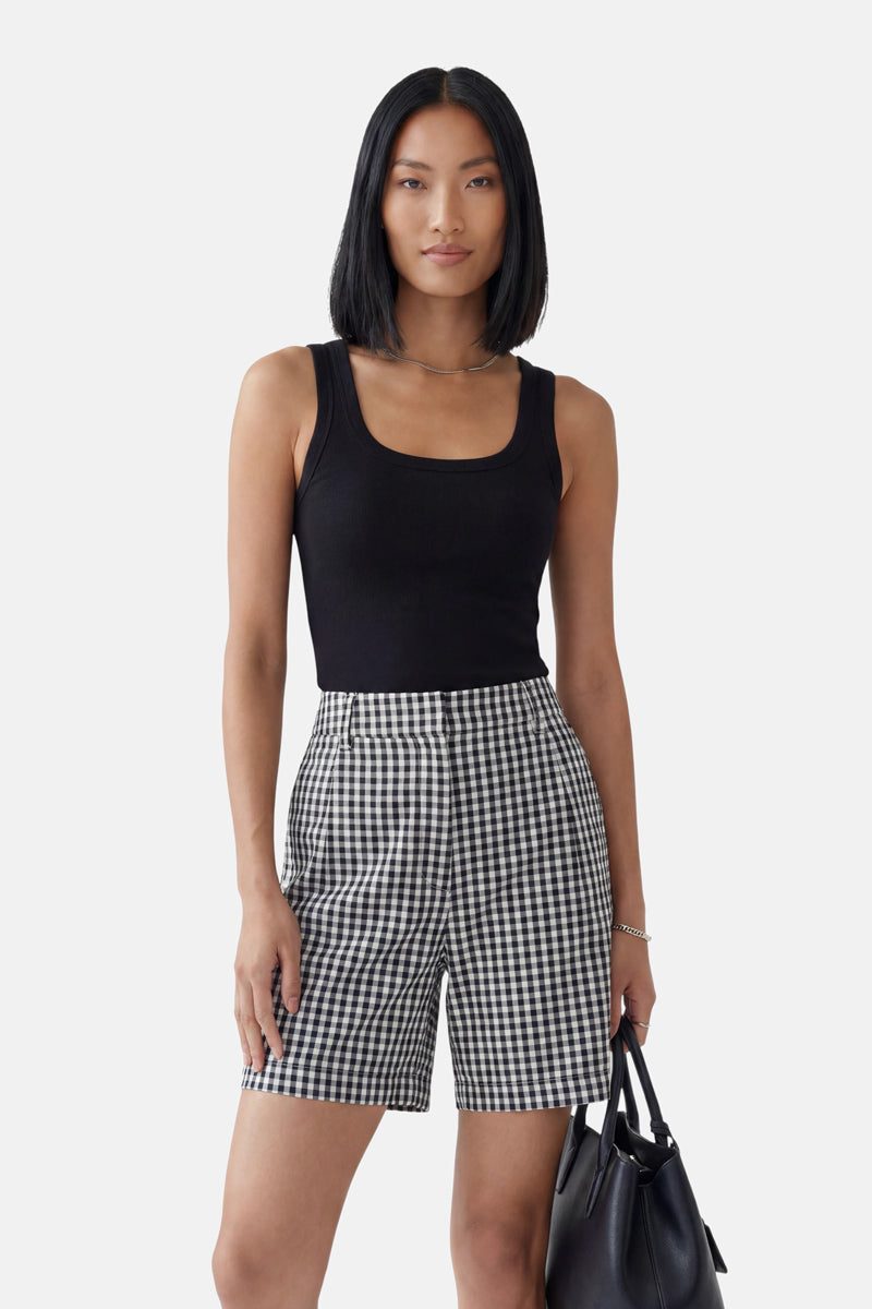 Eine Frau mit glattem schwarzem Haar präsentiert ein schwarzes „Princess Goes Hollywood Basic Top“ mit Rundhalsausschnitt und hoch taillierten schwarz-weiß karierten Shorts. Sie hält eine schwarze Handtasche in der Hand und steht selbstbewusst vor einem schlichten hellgrauen Hintergrund.