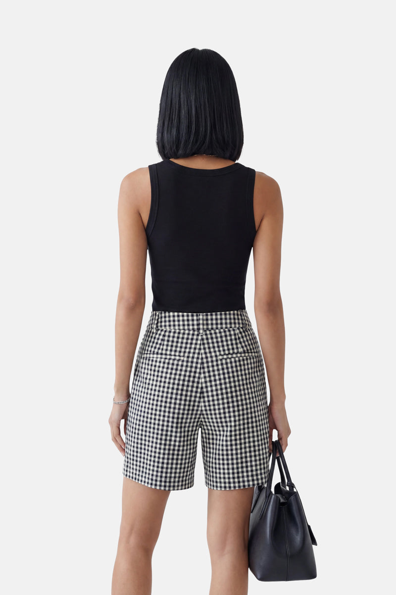 Eine Person mit glattem, schulterlangem schwarzem Haar ist von hinten zu sehen. Sie trägt ein eng anliegendes schwarzes „Princess Goes Hollywood Basic Top“ mit Rundhalsausschnitt und schwarz-weiß karierte Shorts mit hoher Taille. Vor einem schlichten weißen Hintergrund hält sie eine schwarze Handtasche in der Hand.