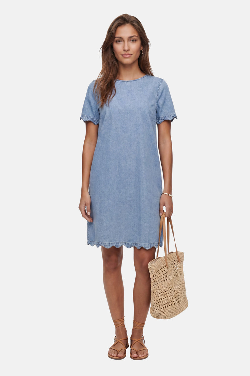 Vor einem weißen Hintergrund steht eine Frau in einem Minikleid aus Jeansstoff der Marke „Princess Goes Hollywood“ mit Wellenkanten. Sie trägt eine geflochtene Strohtasche und hellbraune Schnürsandalen, und ihr offenes braunes Haar umrahmt ihren entspannten Gesichtsausdruck.