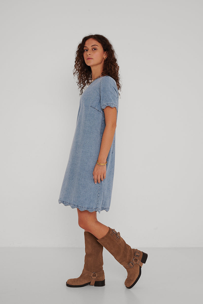 Eine Frau mit lockigem braunem Haar steht seitlich vor einem weißen Hintergrund. Sie trägt ein locker sitzendes, hellblaues Princess Goes Hollywood-Minijeanskleid mit kurzen Ärmeln und braune kniehohe Wildlederstiefel. Sie trägt ein goldenes Armband an ihrem linken Handgelenk.