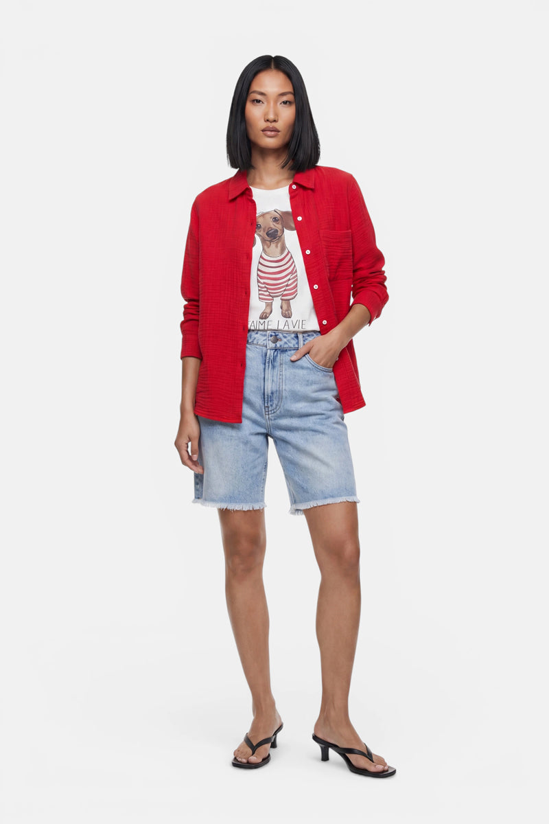 Eine Frau mit glattem schwarzem Haar trägt hellblaue Jeansshorts von „Princess Goes Hollywood“ mit leichter Waschung und ausgefranstem Saum, eine rote Bluse über einem T-Shirt mit Hundemotiv sowie schwarze Sandalen mit Absatz und steht vor einem schlichten weißen Hintergrund.